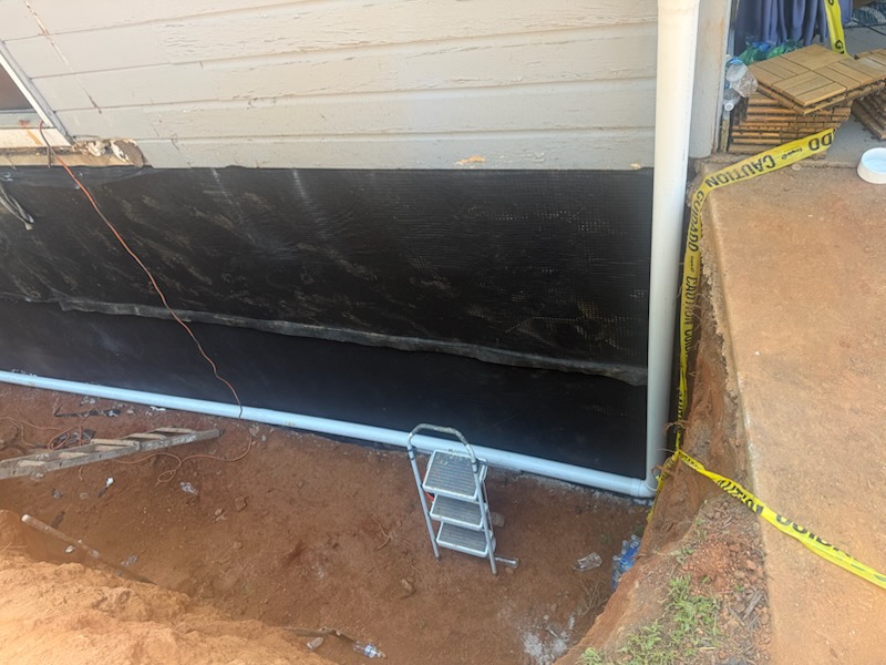 Foundation wall wrapped in black waterproofing membrane with a white drain pipe along the base of the excavation