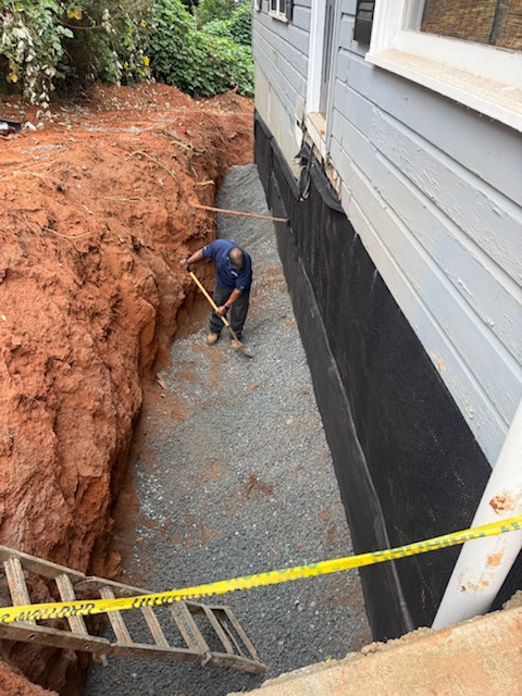 Crew member spreading gravel in an excavated trench beside a foundation wall covered with black waterproofing membrane