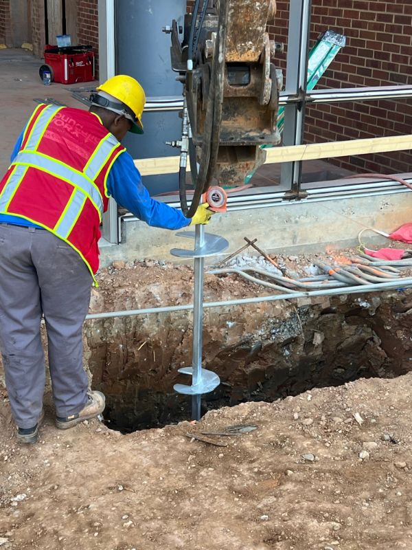 Crew member guiding a helical pile installation beside a commercial building