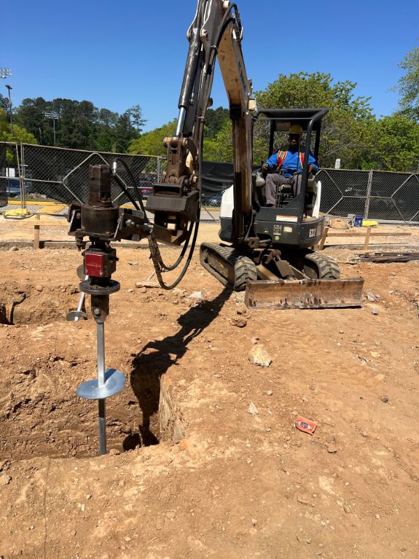 Compact excavator with a hydraulic drive head installing a helical pile on a fenced job site